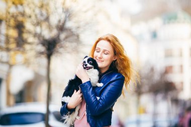 Genç redhaired beyaz bir kadın yüzündeki çiller ile tutar ve öper, hugs, siyah ve beyaz tüylü köpek Chihuahua cins seviyor. Baharda ayakta dışında mavi deri ceket giymiş kız