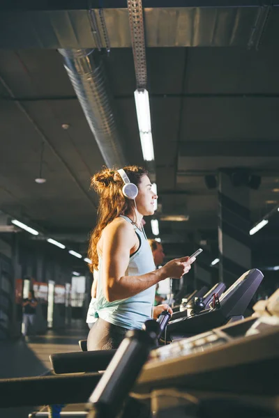 Tema deporte y música. Una hermosa mujer caucásica corriendo en el gimnasio en la cinta de correr. En la cabeza grandes auriculares blancos, la chica escucha música durante un entrenamiento cardiovascular para bajar de peso y utiliza el teléfono en la mano — Foto de Stock