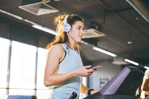 Theme sport and music. A beautiful Caucasian woman running in gym on treadmill. On head big white headphones, the girl listens to music during a cardio workout for weight loss and uses phone in hand
