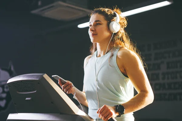 Tema deporte y música. Una hermosa mujer caucásica corriendo en el gimnasio en la cinta de correr. En la cabeza grandes auriculares blancos, la chica escucha música durante un entrenamiento cardiovascular para bajar de peso y utiliza el teléfono en la mano — Foto de Stock