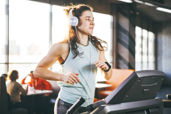 El tema es deporte y música. Una hermosa mujer inflada corre en el gimnasio en una cinta de correr. En su cabeza hay grandes auriculares blancos, la chica escucha música durante un entrenamiento cardiovascular para bajar de peso — Foto de Stock