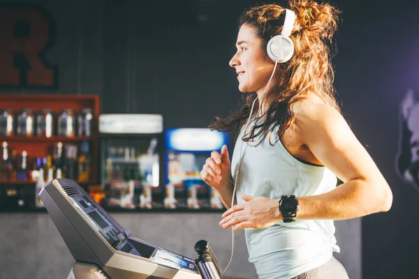 El tema es deporte y música. Una hermosa mujer inflada corre en el gimnasio en una cinta de correr. En su cabeza hay grandes auriculares blancos, la chica escucha música durante un entrenamiento cardiovascular para bajar de peso — Foto de Stock