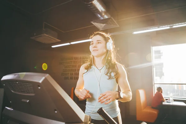 Theme is sport and music. A beautiful inflated woman runs in the gym on a treadmill. On her head are big white headphones, the girl listens to music during a cardio workout for weight loss