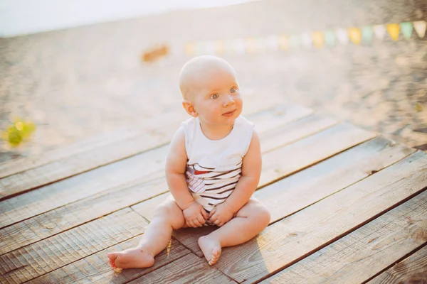 Enfant garçon d'un an blond assis sur un quai en bois, une jetée en vêtements rayés, un complexe près de l'étang sur une plage de sable sur un fond de rivière en été au coucher du soleil de la journée — Photo