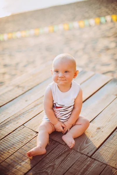 Один год блондин сидит на деревянном причале, на пирсе в полосатой одежде, рядом с прудом на песчаном пляже на фоне реки летом на закате дня — стоковое фото