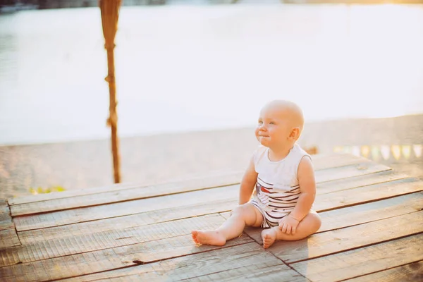 夏の日の夕暮れ時のウッドデッキは、ストライプの服で桟橋、川を背景に砂浜で池の近くの化合物に座っている少年 1 年金髪の子 — ストック写真
