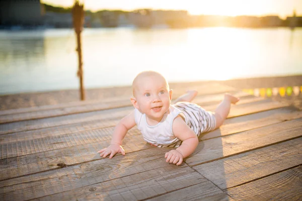 Ребенок мальчик, годовалый блондин лежит на животе на деревянном причале, пирс в полосатой одежде, усадьба возле пруда на песчаном пляже на фоне реки летом на закате дня — стоковое фото