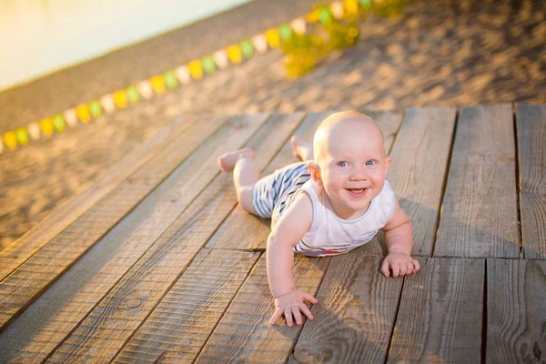 Un niño un niño, un año de edad, hombre rubio se acuesta sobre su estómago en el muelle de madera, muelle con ropa a rayas, compuesto cerca de estanque en la playa de arena contra el fondo del río en el verano al atardecer del día —  Fotos de Stock