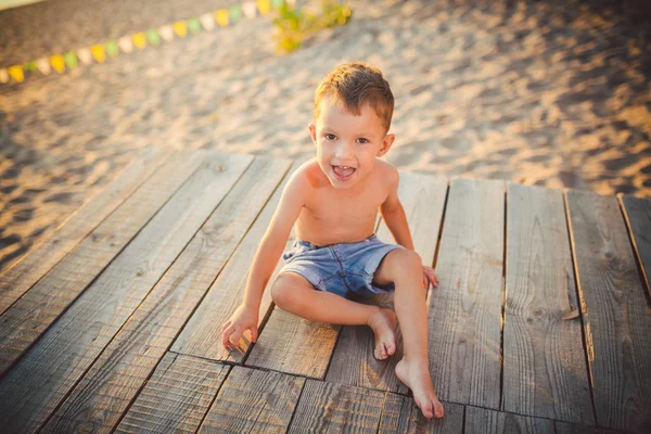 El tema es vacaciones en la playa de verano y niños. Un pequeño niño caucásico se sienta de costado en un muelle de madera y admira la vista de la playa de arena y el estanque, el río. Con piernas desnudas en pantalones cortos de mezclilla azul —  Fotos de Stock
