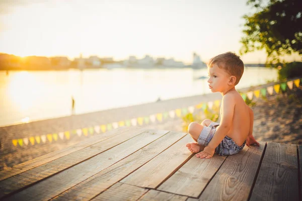 Тема детские и летние пляжные каникулы. Маленький кавказский мальчик сидит боком на деревянном пирсе и любуется видом на песчаный пляж и пруд, реку. С голыми ногами в джинсовых шортах — стоковое фото