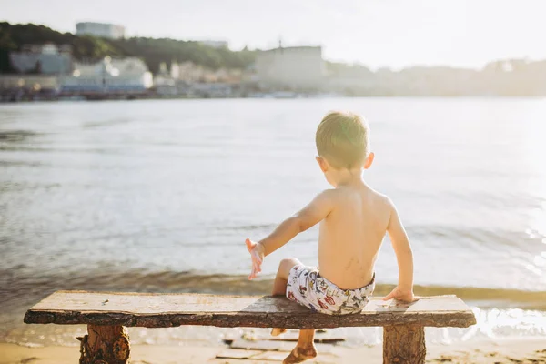 Niño sentado en un banco de madera con la espalda en la playa cerca del agua y mostrando su mano hacia adelante en el telón de fondo de la ciudad de Kiev —  Fotos de Stock