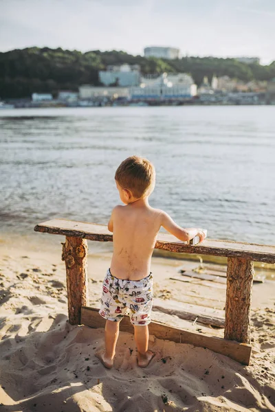 Тема детские и летние пляжные каникулы. Маленький кавказский мальчик забирается на деревянную скамейку на берегу реки в солнечный летний день на песчаном пляже с видом на город — стоковое фото