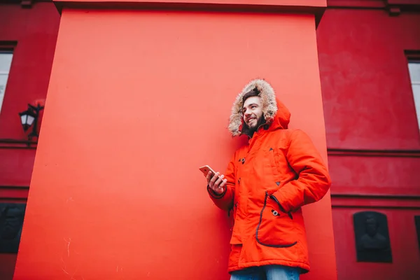 Jovem estudante bonito com sorriso de dente e barba fica no fundo da parede vermelha em casaco de inverno vermelho brilhante com capuz com pele, usa, mantém o telefone móvel com a mão no bolso. Inverno tempo frio — Fotografia de Stock