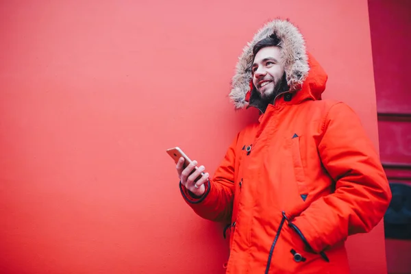 Handsome young male student with toothy smile and beard stands on red wall background in bright red winter jacket with hood with fur, Uses, holds mobile phone with hand in pocket. Winter cold weather — Stock Photo, Image