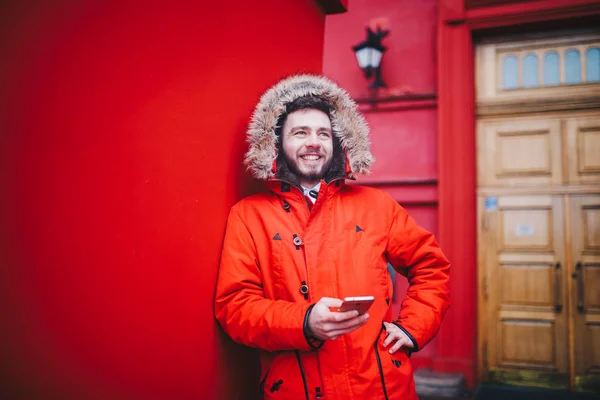 Jovem estudante bonito com sorriso de dente e barba fica no fundo da parede vermelha em casaco de inverno vermelho brilhante com capuz com pele, usa, mantém o telefone móvel com a mão no bolso. Inverno tempo frio — Fotografia de Stock