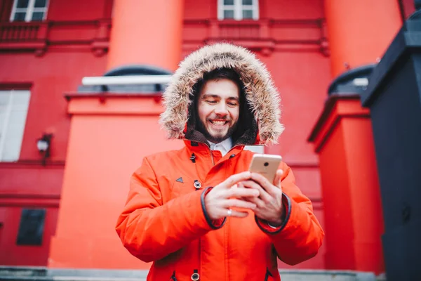 こぼれるような笑顔とひげのハンサムな若い男子学生が赤い壁背景、毛皮のフード、携帯電話の画面で使用する指と赤いジャケットの教育機関のファサードの上に立つ — ストック写真