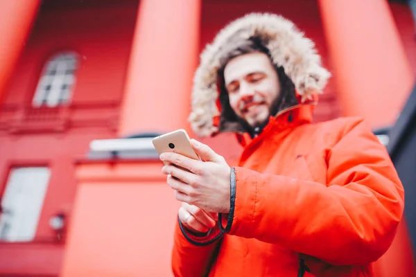 Przystojny młody mężczyzna student z Uśmiech zębiczny i broda stoi na tle czerwone ściany, fasady instytucji edukacyjnych w Czerwona zimowa kurtka kaptur z futerkiem, używa palec na ekranie telefonu komórkowego — Zdjęcie stockowe