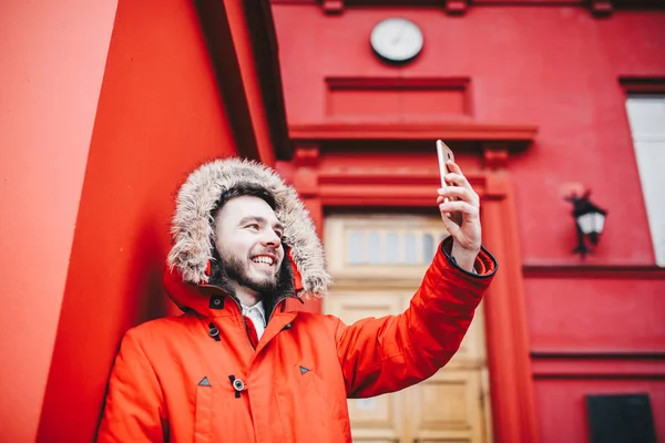 Jovem estudante bonito com sorriso e barba fica no fundo parede vermelha, fachada da instituição de ensino em casaco de inverno vermelho com um capuz com pele no inverno. Usa um telemóvel, faz selfie — Fotografia de Stock