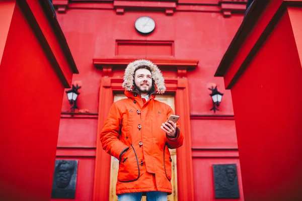 Handsome young male student with toothy smile and beard stands on red wall background in bright red winter jacket with hood with fur, Uses, holds mobile phone with hand in pocket. Winter cold weather — Stock Photo, Image