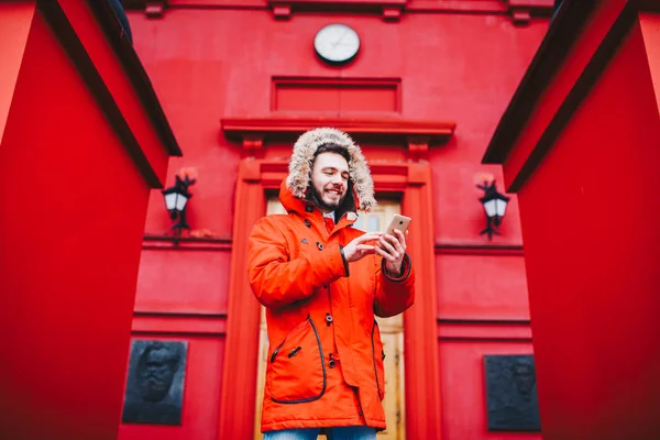 Jovem estudante bonito com sorriso de dente e barba fica no fundo da parede vermelha, fachada da instituição de ensino em casaco de inverno vermelho com capuz com pele, usa o dedo na tela do telefone móvel — Fotografia de Stock