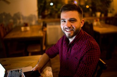 Genç yakışıklı bir beyaz adam sakal ve kırmızı Kareli gömlekli dişlek gülümseme ile tahta masada oturan dizüstü arkasında çalışıyor. Mobil telefon elinde tutan kullanır. Akşamları coffee shop