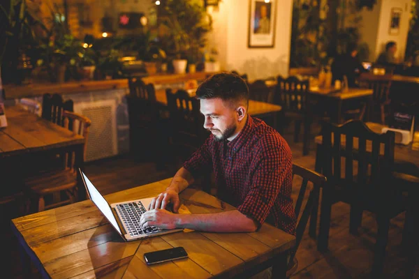 A young handsome Caucasian man with beard and toothy smile in a red checkered shirt is working behind a gray laptop sitting at a wooden table. Hands on the keyboard. In the evening at the coffee shop