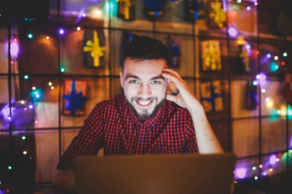 Een jonge knappe blanke man met baard en toothy glimlach in een rode geruite shirt werkt achter een grijze laptop zitten op een houten tafel. Handen op het toetsenbord. In de avond bij de coffeeshop — Stockfoto
