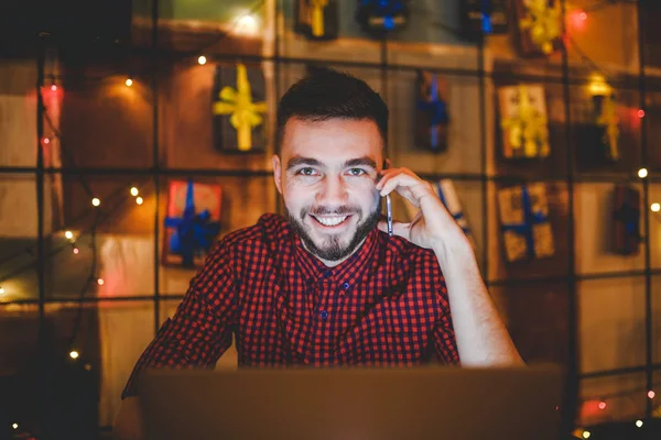 Jonge knappe blanke man met baard en toothy glimlach in rood shirt werken achter de laptop, handen op toetsenbord zitten aan houten tafel. Toepassingen doet een beroep op de mobiele telefoon. In de avond bij de koffieshop — Stockfoto