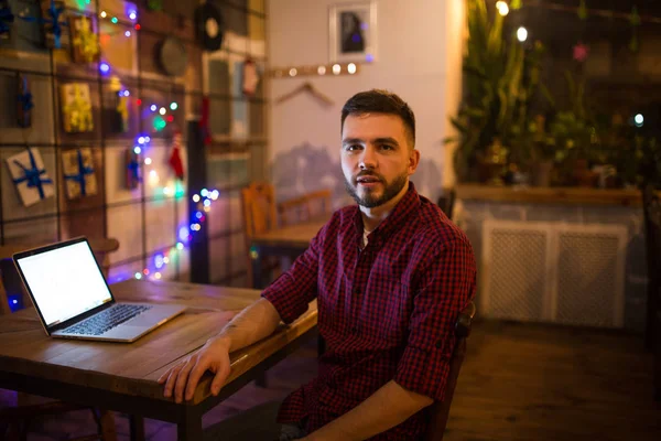 Retrato de un joven guapo hombre caucásico con barba y sonrisa dentada en una camisa roja a cuadros sentada junto a un portátil en una mesa de madera. Por la noche en la cafetería —  Fotos de Stock