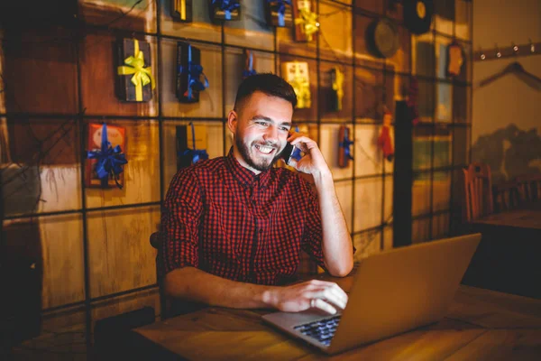 Joven hombre caucásico guapo con barba y sonrisa dentada en camisa roja funciona detrás de la computadora portátil, las manos en el teclado sentado en la mesa de madera. Utiliza llamadas en el teléfono móvil. Por la noche en la cafetería — Foto de Stock