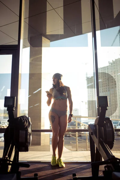 En vacker sexig kaukasisk kvinna står i full tillväxt nära ett fönster med solljus i gymmet. Håller i sin hand, använder en mobiltelefon och sport klocka. Klädd i grå sportkläder och en hatt — Stockfoto