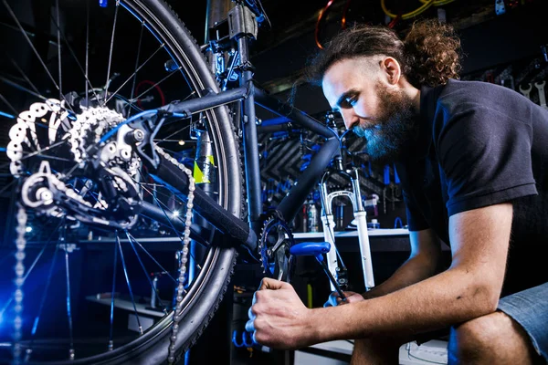 Молодой стильный бородатый, с длинными волосами кавказский механик-мужчина в велосипедной мастерской использует инструмент для ремонта комплектов Cranksets — стоковое фото