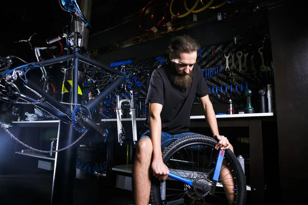 Молодой стильный бородатый, с длинными волосами кавказский механик-мужчина в веломастерской использует инструмент для ремонта кассеты на заднем колесе велосипеда — стоковое фото