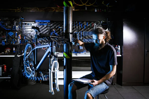 Joven hombre barbudo con estilo con pelo largo hombre caucásico empresario empresario trabajador en taller de bicicletas utiliza cuaderno para escribir notas sentado junto a la bicicleta en el fondo de la herramienta dentro de la tienda —  Fotos de Stock