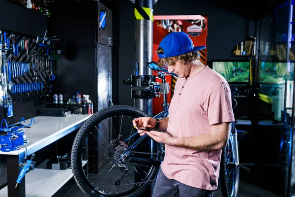 A young, stylish redheaded male small business owner selling and repairing a bike is wearing a blue cap and a pink jersey is using a mobile phone while standing in a bike shop