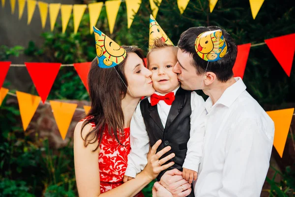 children birthday party. Family father and mother hold son of one year of birth in arms and kiss cheekin the background of park and festive decor with colored ornaments. On heads of hats, funny caps