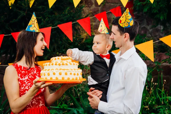 Utsätta barn födelsedagsfest, mat och godis. En ung familj firar ett år son. Pappa håller en stor tårta, Mamma håller en bebis i famnen. Baby smaker finger grädde på tårtan med tårta — Stockfoto