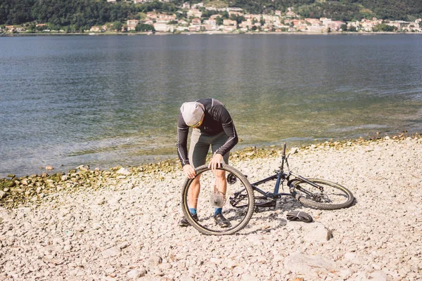 Naprawa rowerów. Człowiek naprawiający rower górski. Rowerzysta w tarapatach, wypadek na tylnym kole. Człowiek naprawia rower w pobliżu jeziora we Włoszech tło gór. rowerzysta naprawiający koło rowerowe na zewnątrz — Zdjęcie stockowe