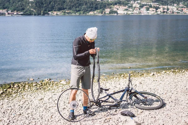 Naprawa rowerów. Człowiek naprawiający rower górski. Rowerzysta w tarapatach, wypadek na tylnym kole. Człowiek naprawia rower w pobliżu jeziora we Włoszech tło gór. rowerzysta naprawiający koło rowerowe na zewnątrz — Zdjęcie stockowe