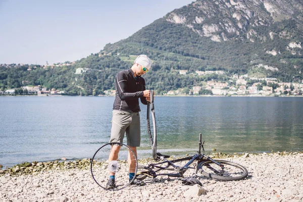 自転車修理。マウンテンバイクを修理する男。事故のトラブル後輪ケースにサイクリストの男。男はイタリアの背景の山の湖の近くの自転車を修正します。自転車を屋外で修理するサイクリスト — ストック写真