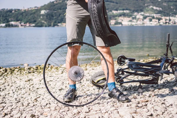 自転車修理。マウンテンバイクを修理する男。事故のトラブル後輪ケースにサイクリストの男。男はイタリアの背景の山の湖の近くの自転車を修正します。自転車を屋外で修理するサイクリスト — ストック写真