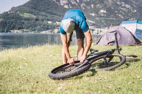 Naprawa rowerów. Człowiek naprawiający rower górski. Rowerzysta w tarapatach, wypadek na tylnym kole. Człowiek naprawia rower w pobliżu jeziora we Włoszech tło gór. rowerzysta naprawiający koło rowerowe na zewnątrz — Zdjęcie stockowe