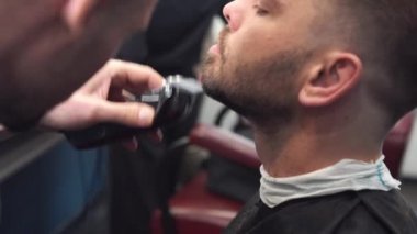 Berber müşterilerin sakallarını sandalyede tıraş eder. Sakal tıraşı. Elektrikli jiletle sakal tıraşı olan bir berber. Gerçek bir erkeğin tımarı. Sakallı genç bir adamın kuaförde sakal tıraşı olması.