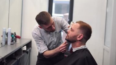 Berber müşterilerin sakallarını sandalyede tıraş eder. Sakal tıraşı. Elektrikli jiletle sakal tıraşı olan bir berber. Gerçek bir erkeğin tımarı. Sakallı genç bir adamın kuaförde sakal tıraşı olması.