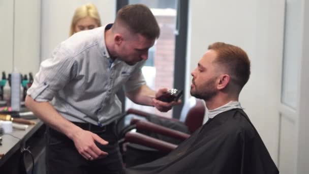 Barber afeita la barba de los clientes en una silla. Corte de pelo de barba. Peluquero para afeitarse la barba con una navaja eléctrica. Aseo de hombre de verdad. Vista lateral del joven barbudo que se corta la barba en la peluquería — Vídeos de Stock