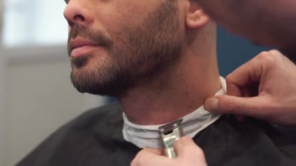 Barberaren rakar skägget på en stol. Skäggig frisyr. Frisör att raka ett skägg med en elektrisk rakhyvel. Omhändertagande av en riktig man. Sidovy av ung skäggig man som får skägg frisyr hos frisör — Stockvideo