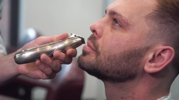 O barbeiro rapa a barba dos clientes numa cadeira. Corte de cabelo de barba. Barbeiro para barbear uma barba com uma navalha elétrica. Preparação de um homem a sério. Vista lateral do jovem barbudo recebendo corte de cabelo barba no cabeleireiro — Vídeo de Stock