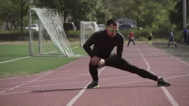 2 de março de 2019. Ucrânia, Kiev. Tema esporte e saúde. Homem caucasiano jovem faz exercício de aquecimento alongamento aquecendo os músculos antes de treinar em corrida em uma esteira de pista de estádio da cidade — Vídeo de Stock