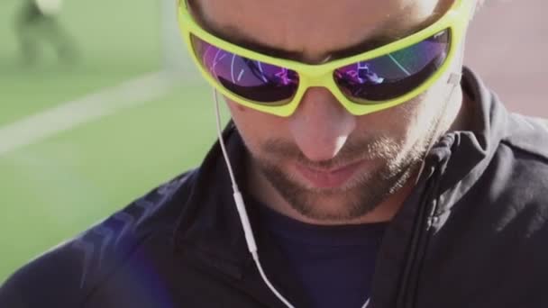 Tema deporte y salud. Un joven caucásico usa un smartphone smarfton para escuchar música en los auriculares. Lista de reproducción de tecnología y aplicaciones móviles en la cinta de correr del estadio de la ciudad — Vídeo de stock