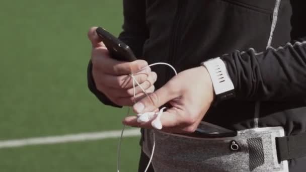 Tema deporte y salud. Un joven caucásico usa un smartphone smarfton para escuchar música en los auriculares. Lista de reproducción de tecnología y aplicaciones móviles en la cinta de correr del estadio de la ciudad — Vídeo de stock